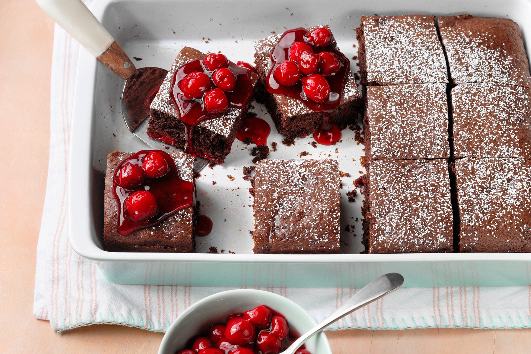 A rectangular baking dish with chocolate brownies topped with cherry sauce and powdered sugar. Some brownies are cut into squares. A serving spatula and a bowl with extra cherry sauce and a spoon are beside the dish.