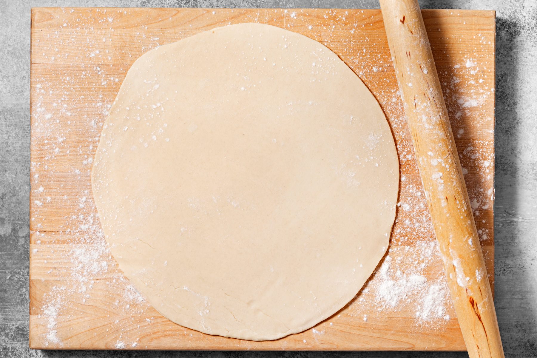 overhead shot of a flat round piece of dough on a wooden cutting board, there is a rolling pin lying on the right side of the board, there are some specks of flour on the board and on the dough, the background is gray.