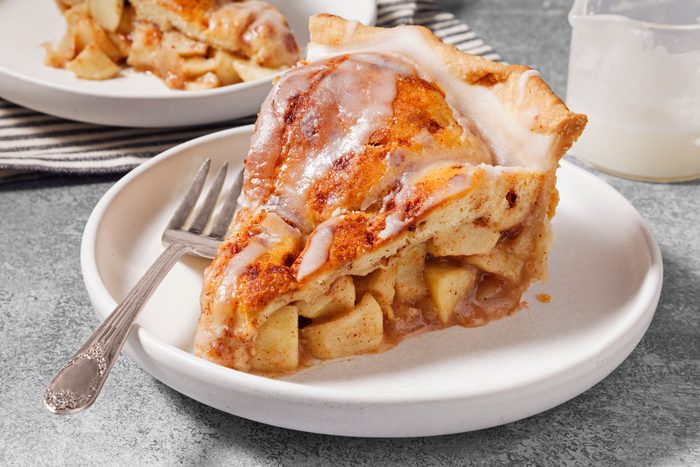 3/4th shot of a slice of apple pie sits on a white plate, with a silver fork placed on the plate, and the handle of the fork is visible; the plate is on a grey textured surface; a blurred view of another slice of apple pie on a white plate can be seen to the left of the main slice; a pitcher of milk is slightly out of focus to the right; the apple pie is topped with a white glaze and cinnamon; the pie crust is a light golden brown