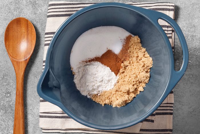overhead shot of a blue mixing bowl with dry ingredients for baking; there is white sugar, brown sugar, flour, and cinnamon in the bowl; the bowl is on top of a white and black striped kitchen towel; a wooden spoon is next to the bowl on the left side of the image; the background is a light gray textured surface