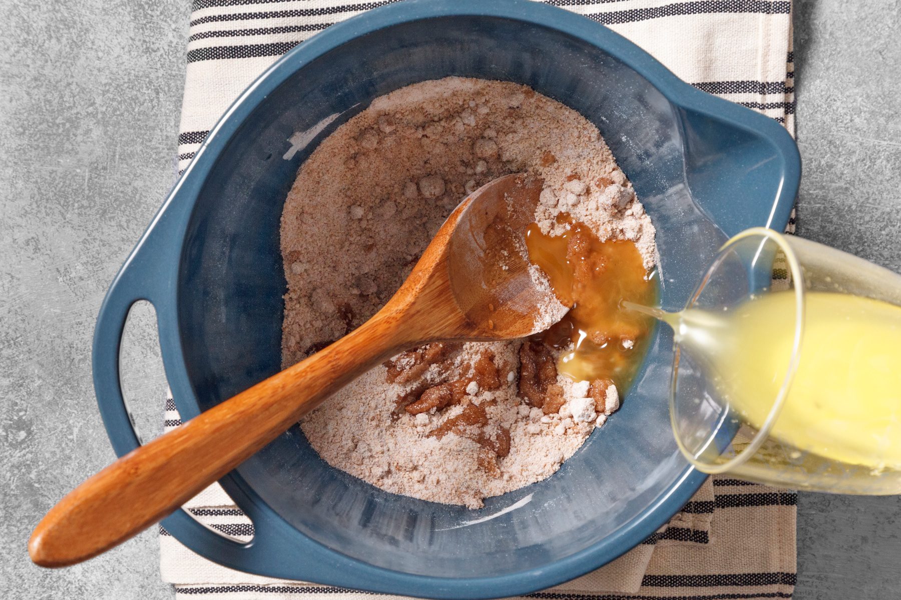 overhead shot of a blue bowl filled with dry ingredients, likely for baking; the bowl is resting on a striped kitchen towel, and there is a wooden spoon in the bowl, which is stirring the dry ingredients, a clear glass with liquid is being poured into the bowl from the right side; the background is a textured grey surface