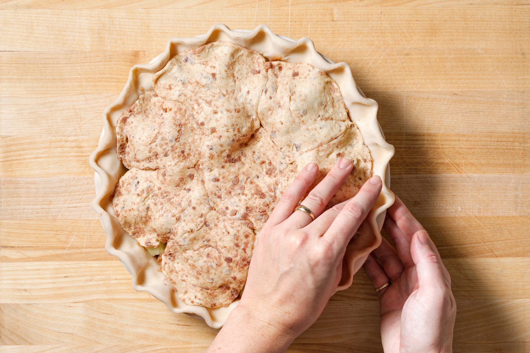 overhead shot of a partially prepared pie resting on a light wooden surface, with a fluted crust neatly pressed along the edges of a pie dish, two hands are visible, one holding the pie dish for support and the other gently adjusting the crust edges to ensure an even shape
