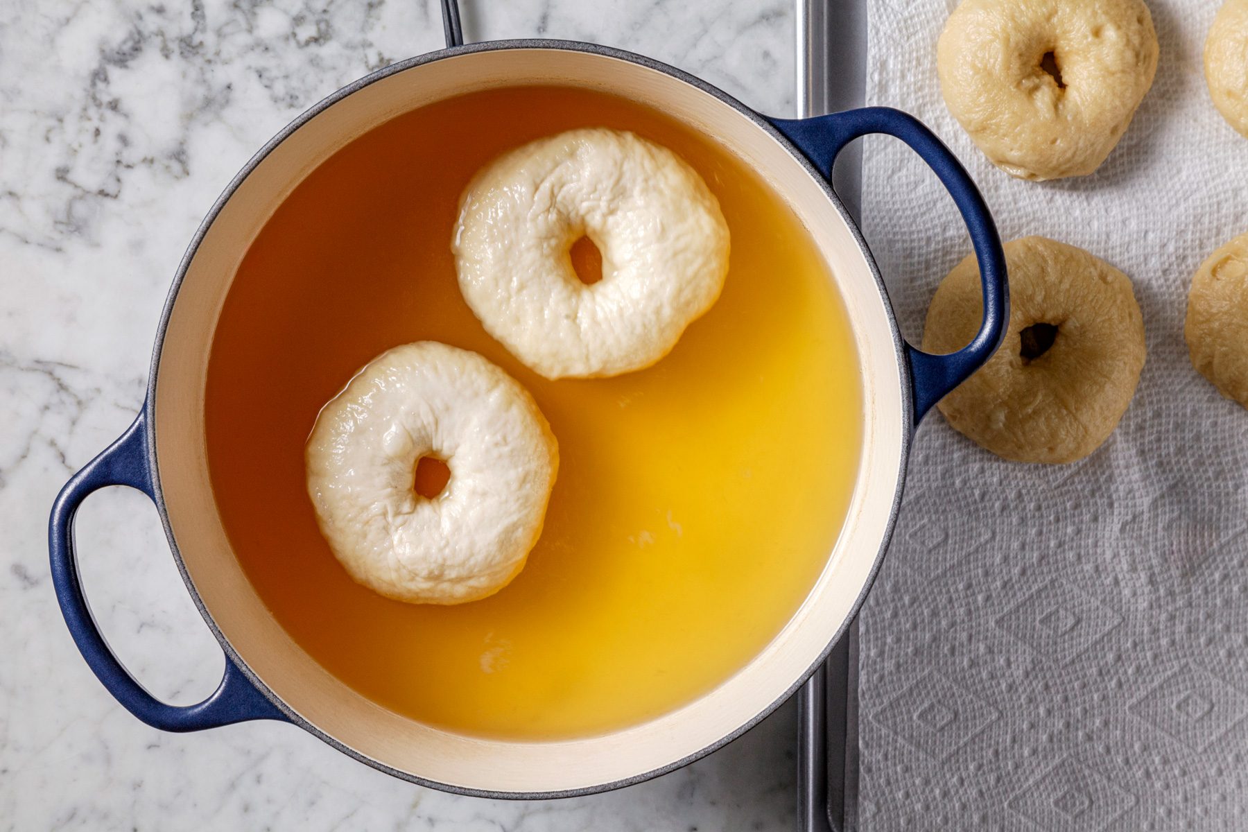 Overhead shot of a Dutch oven filled with water and honey, brought to a boil; adding bagels two at a time, cooking for 45 seconds per side, then removing with a slotted spoon to drain on paper towels, on a marble surface.