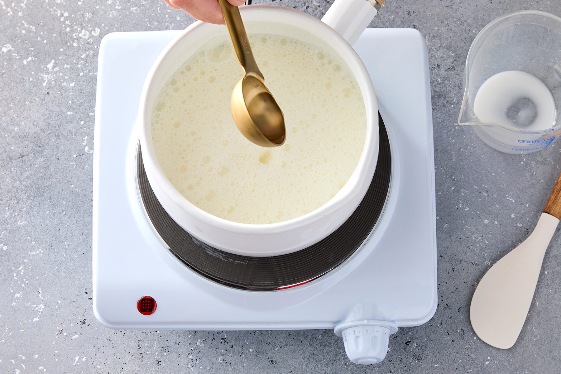 A person stirs white creamy liquid in a pot on an electric burner with a brass spoon. A measuring cup and spatula are nearby on the gray speckled countertop.