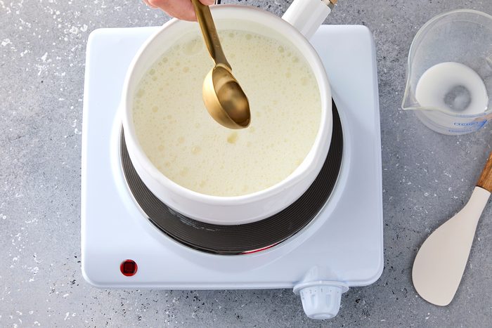 A person stirs white creamy liquid in a pot on an electric burner with a brass spoon. A measuring cup and spatula are nearby on the gray speckled countertop.