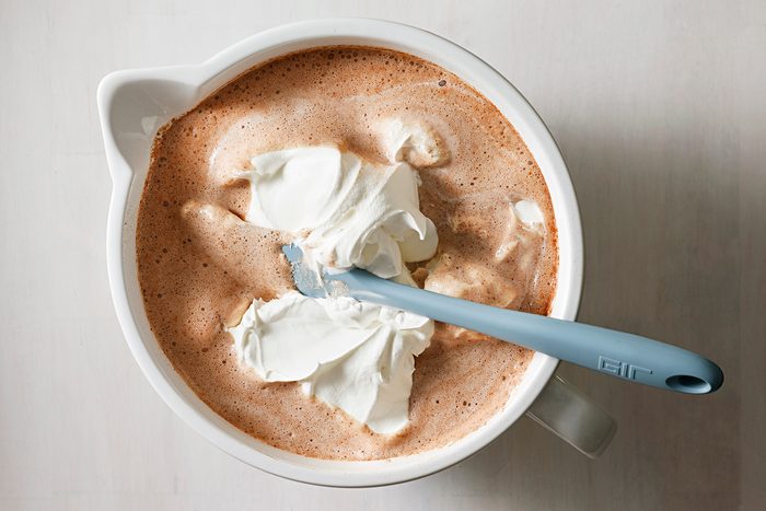 A mixing bowl filled with a creamy cocoa mixture is being stirred with a blue spatula. Fluffy dollops of whipped cream are visible on the surface, creating a swirling pattern.