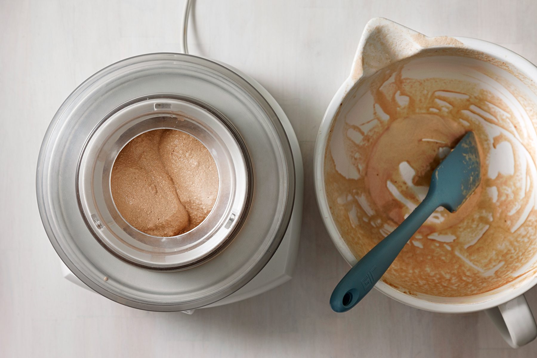 A top-down view of an ice cream maker churning a brown mixture next to a white mixing bowl with leftover batter. A blue spatula rests in the bowl. The scene is set on a light-colored surface.