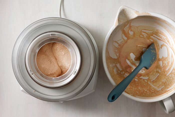 A top-down view of an ice cream maker churning a brown mixture next to a white mixing bowl with leftover batter. A blue spatula rests in the bowl. The scene is set on a light-colored surface.