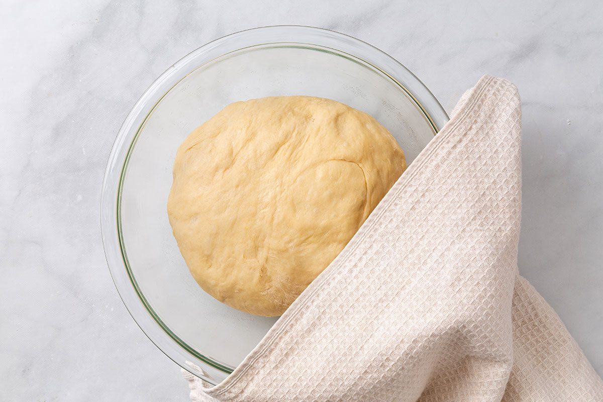 Bread dough in a greased mixing bowl with a dish towel covering for step two of Country White Bread recipe for Taste of Home