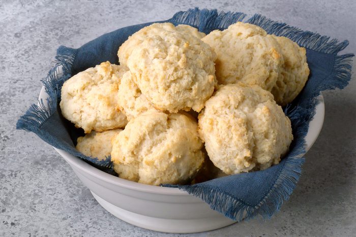 A white bowl filled with golden-brown biscuits sits on a blue cloth. The biscuits have a rough, textured surface and are arranged in a cozy stack. The bowl is placed on a lightly speckled gray surface.