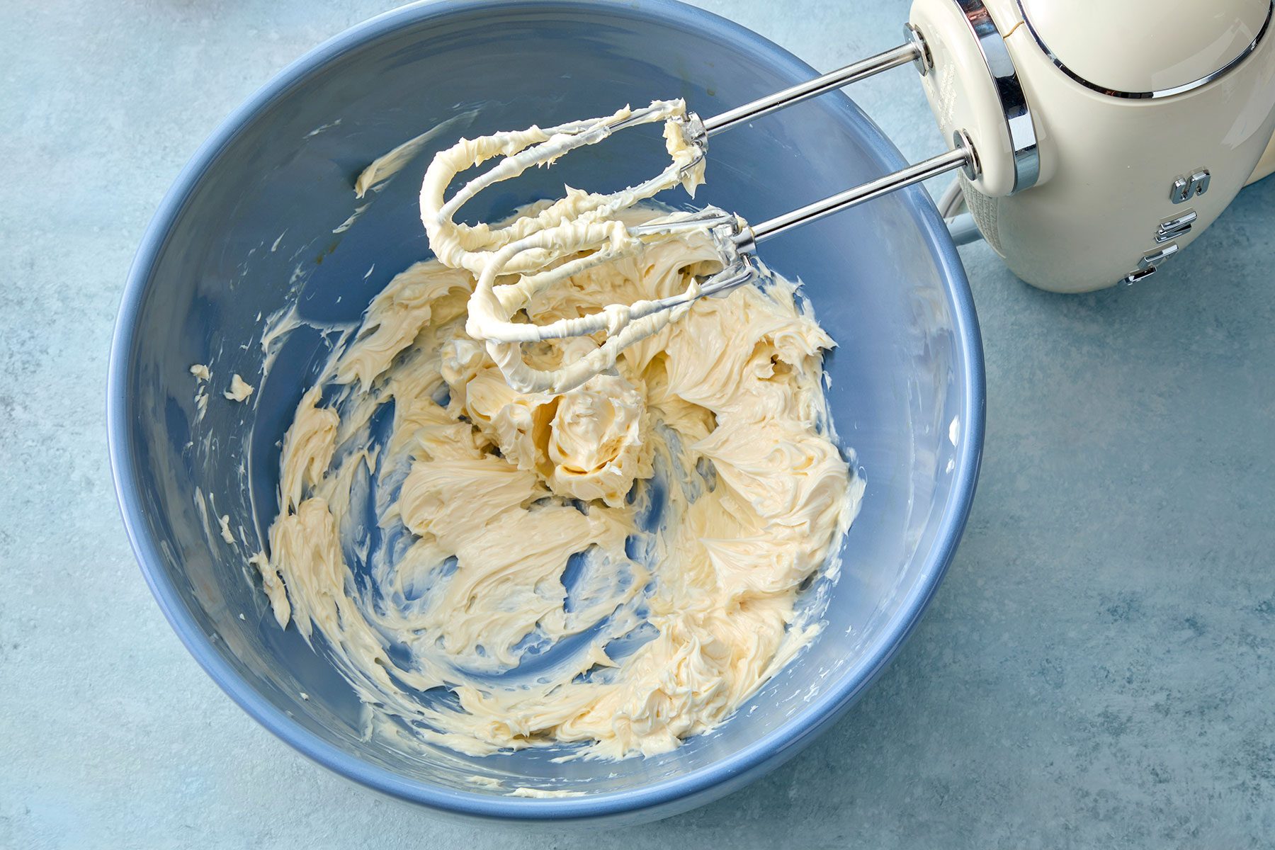 A blue mixing bowl filled with creamy, whipped butter being mixed by an electric hand mixer with beaters covered in the cream. The bowl is placed on a light blue countertop.