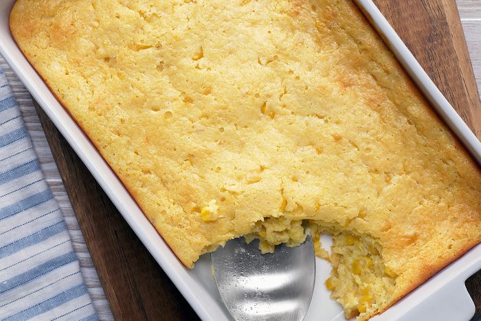 overhead shot of a casserole dish filled with cornbread pudding, it's perfectly browned on top and a silver spatula is used to take a serving from the dish; the dish is sitting on a wooden cutting board with a blue and white striped kitchen towel beside it