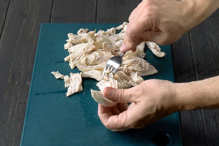 3/4th shot of a person shredding cooked chicken with a fork on a blue cutting board; the chicken is on the cutting board and the person is holding the fork with their right hand and using their left hand to hold the chicken