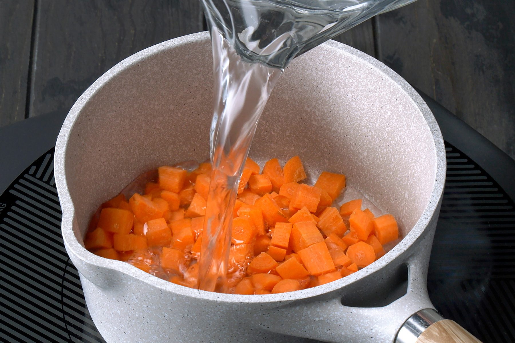 3/4th shot of a pot being filled with water, which is poured from a pitcher, and the pot contains chopped carrots; the pot sits on top of a black burner