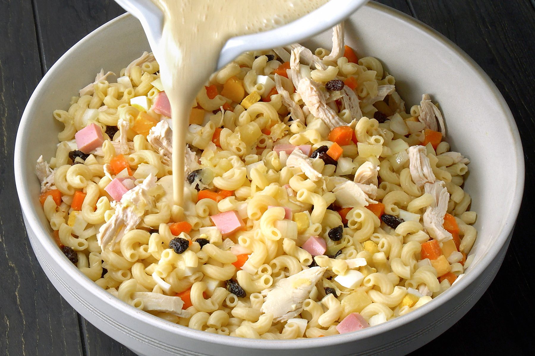 3/4th shot of a white bowl filled with macaroni salad, with a white pitcher pouring a creamy dressing on top; the bowl is resting on a dark wooden table