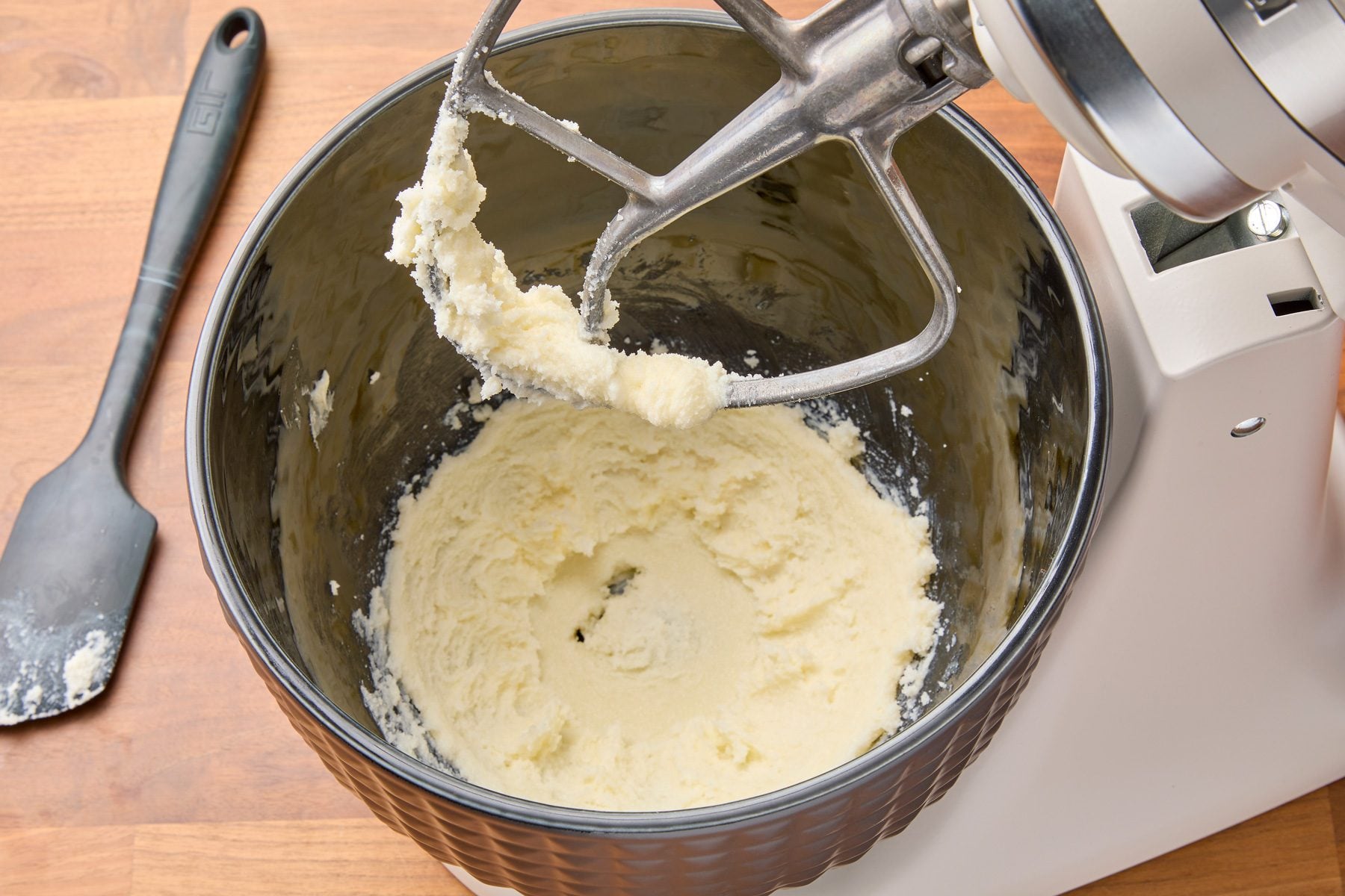 Overhead shot of a large bowl with butter and 2/3 cup sugar being beaten at medium speed until fluffy. A spatula is placed on the wooden countertop to the left of the mixer.