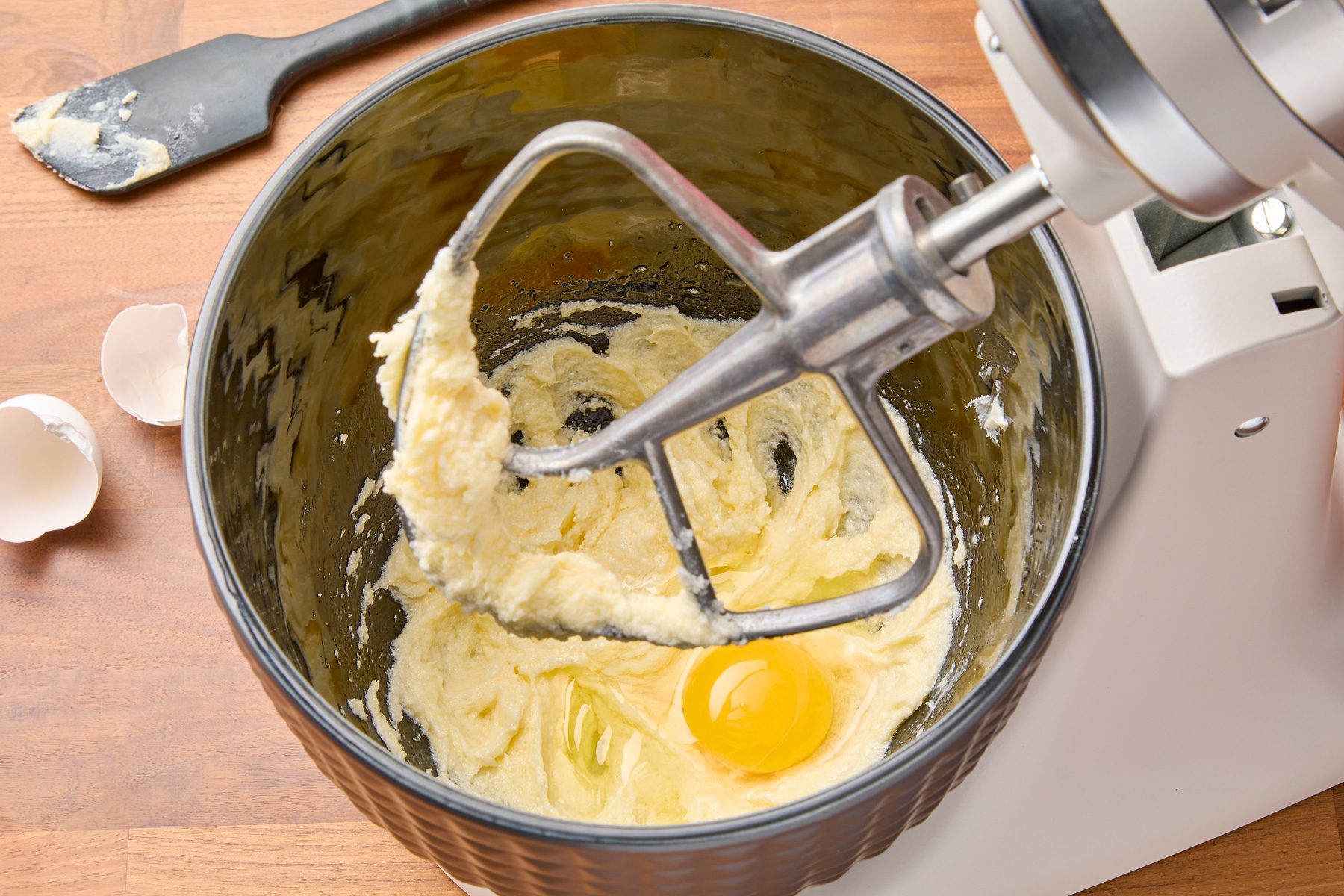 Overhead shot of a stand mixer with a paddle attachment mixing a bowl of batter. The batter contains a single egg yolk in the center, surrounded by white batter, with a bit of batter stuck to the paddle. A spatula is positioned to the left of the bowl, and a wooden table is visible beneath the mixer and spatula. Partially visible eggshells are in the bottom left corner of the image.