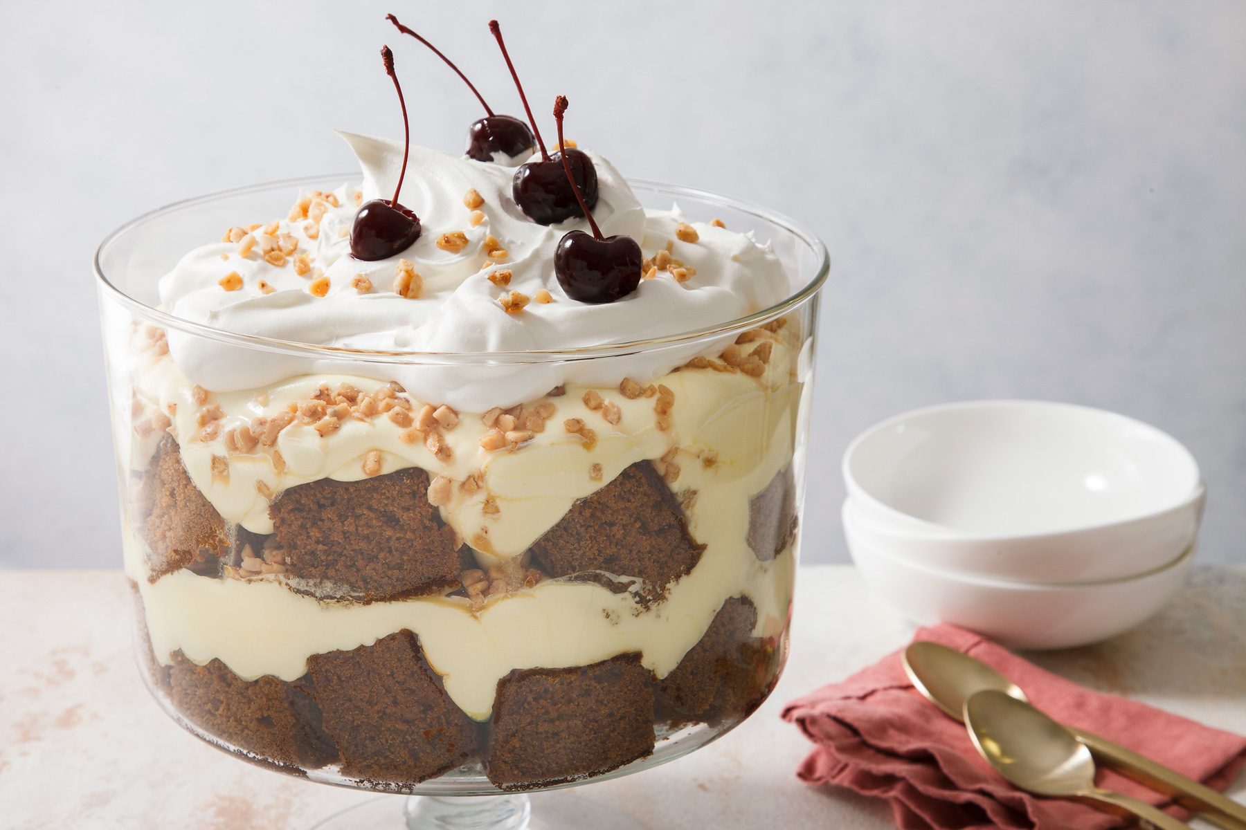 Close shot of Gingerbread Trifle; garnish with cherries; spoons; napkin; bowls; marble surface.