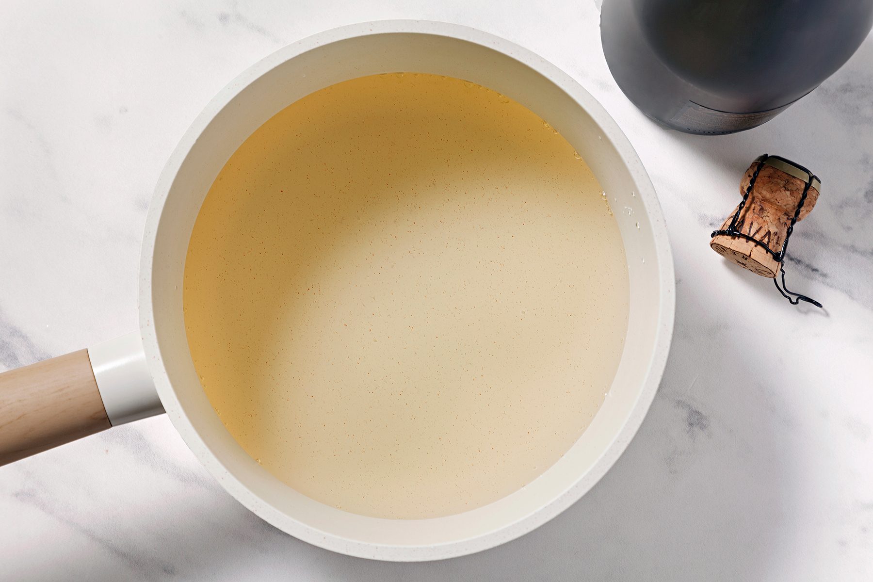 A white saucepan filled with a light yellow creamy sauce on a marble countertop. A bottle and a cork are nearby.