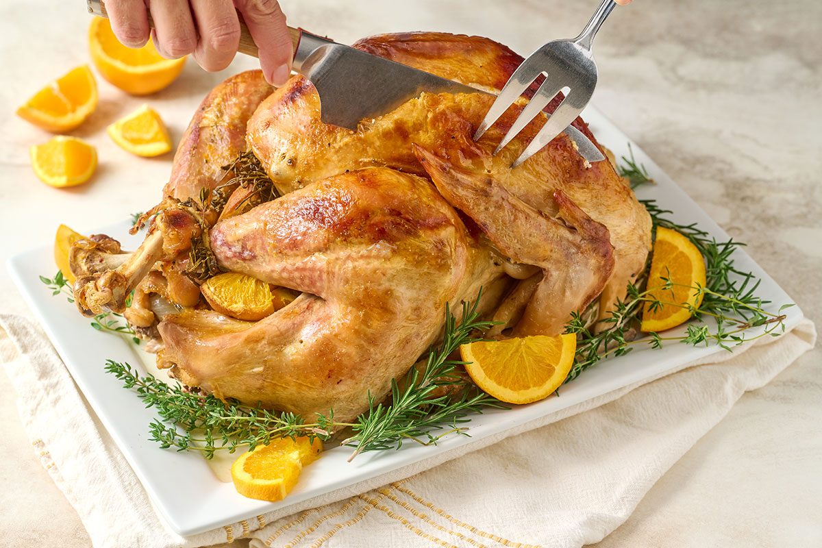 Close-up horizontal shot of the Taste of Home Orange Turkey, getting carved and highlighting the golden, crispy skin and fresh herb garnish.