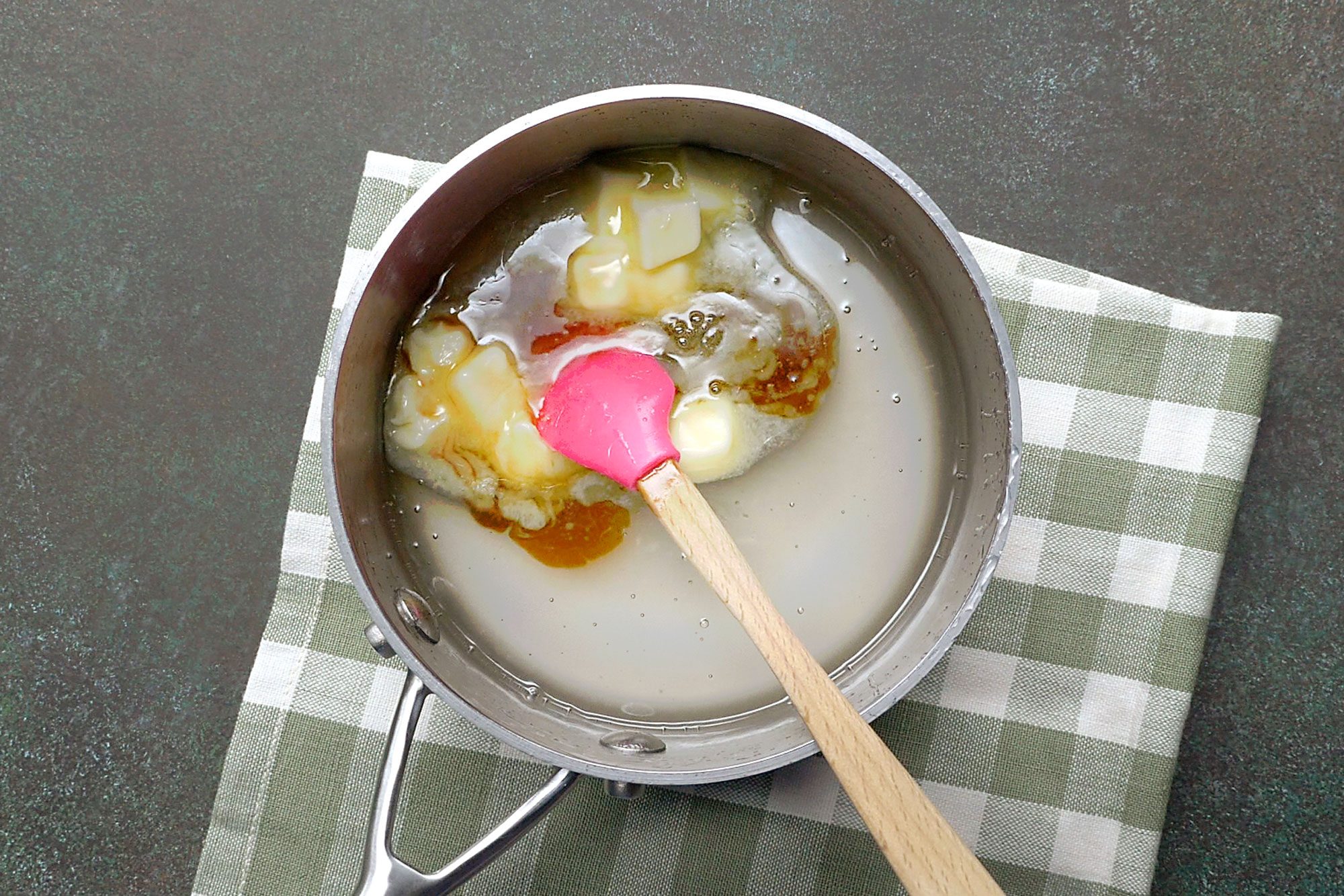 Overhead shot of remove from the heat; add butter; vanilla extract and food coloring; stir until butter is melted; spatula; napkin; green marble surface;
