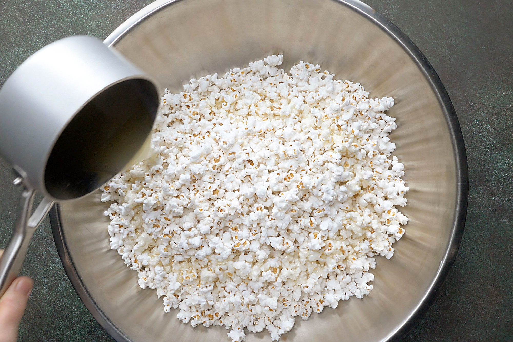Overhead shot of place popcorn in a large baking pan; keep warm in a 200 degree oven; Immediately pour mixture over popcorn and stir until evenly coated; green marble surface;