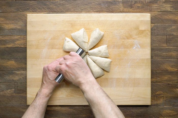 Hands holding a tool, cutting a round piece of dough into equal triangular sections on a wooden cutting board.