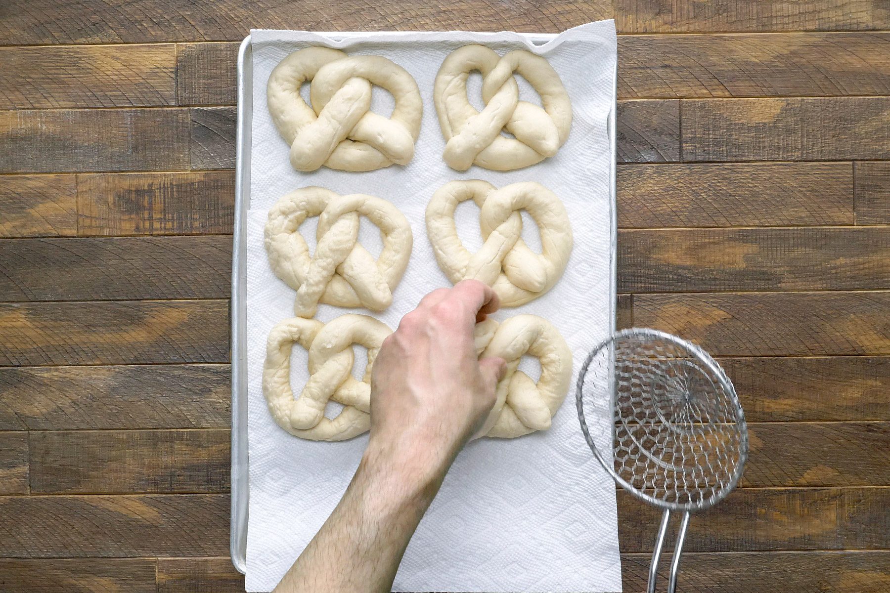 A hand sprinkling salt on six unbaked pretzels arranged on a parchment-lined baking sheet. A metal skimmer rests beside the tray on a wooden surface.