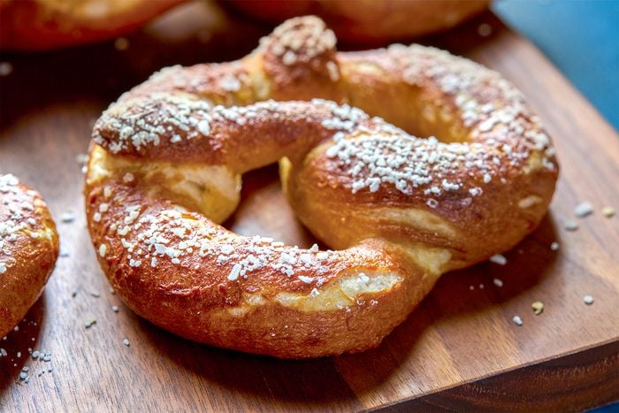 Close-up of a golden-brown pretzel sprinkled with coarse salt, resting on a wooden board. The pretzel's surface is textured and slightly glossy, with a twisted, knot-like shape that is traditional for this type of baked good.