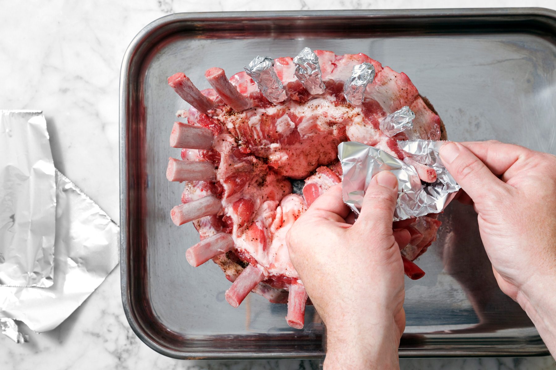 Overhead shot of place roast in a shallow roasting pan; cover rib ends with aluminum foil to keep from burning; pour chicken broth into roasting pan; bake uncovered for 1-3/4 hours; marble surface.