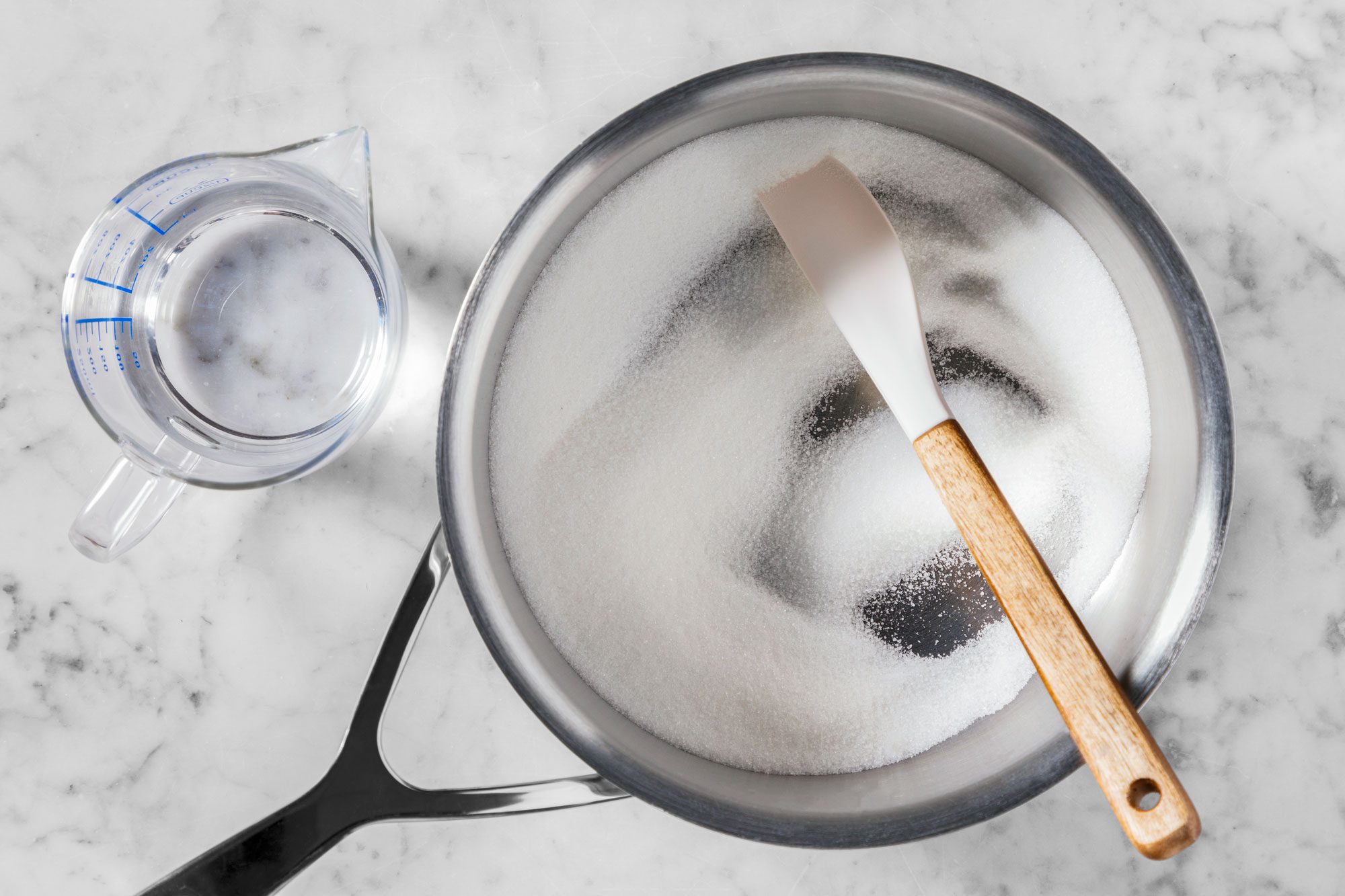 A marble countertop holds a saucepan filled with sugar and a wooden spatula. Nearby is a clear measuring cup with water.