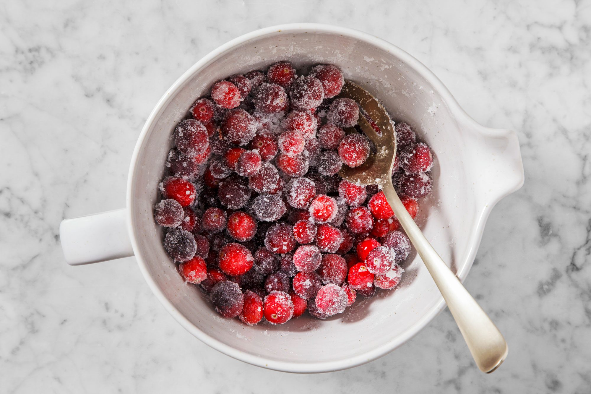 A bowl of sugared cranberries with a silver spoon on a marble surface. The cranberries are coated with a layer of sugar, giving them a frosty appearance.