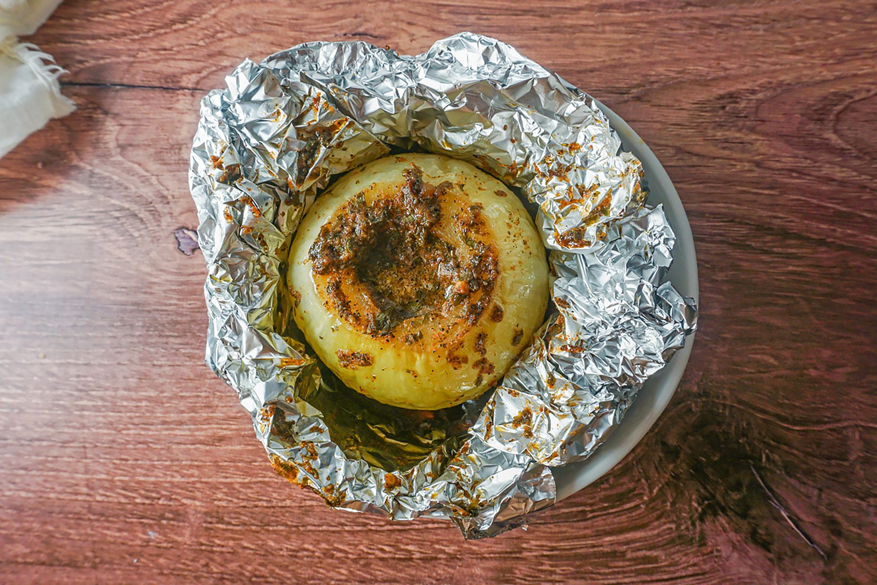 A baked onion wrapped in aluminum foil, partially unwrapped on a small plate. The onion is roasted with a crispy top and seasoned with herbs, set on a wooden table.