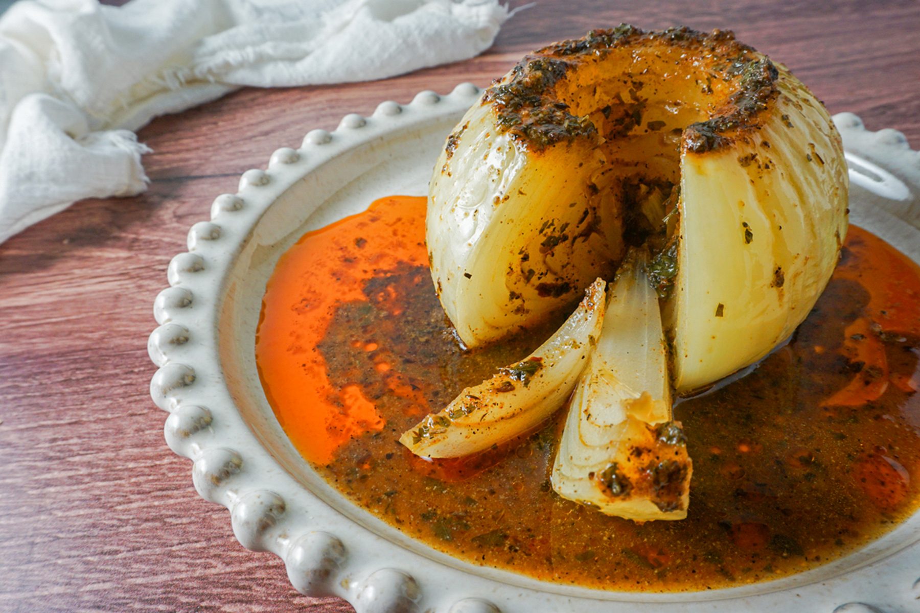 A roasted whole onion served on a decorative white plate, with a rich reddish-orange sauce pooling around it. The onion is cut partially open, showing a seasoned interior. A white cloth is in the background on a wooden surface.