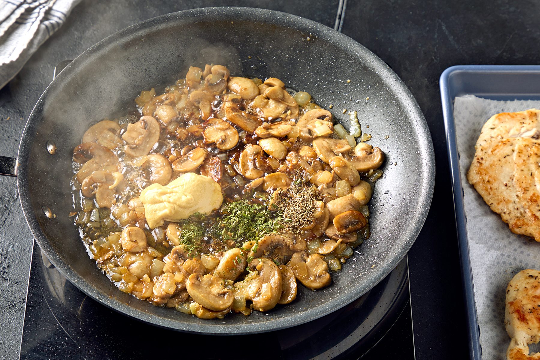 A skillet on a stovetop containing sautéed mushrooms with herbs and a dollop of mustard, emitting steam. On the right, partially visible, is a piece of cooked chicken on a tray lined with paper towels.