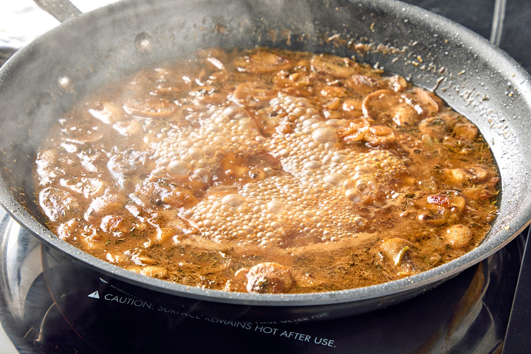 A skillet on a stovetop contains a simmering brown sauce with mushrooms and visible steam, suggesting the mixture is being cooked.