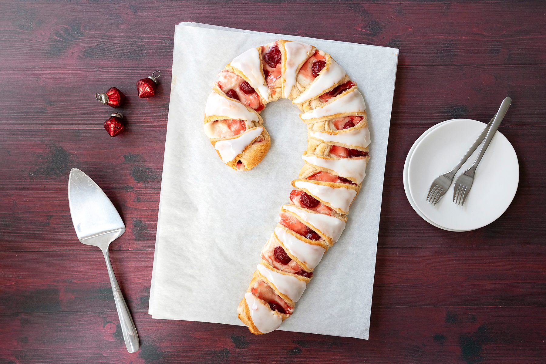A pastry shaped like a candy cane, drizzled with white icing and topped with red fruit, sits on parchment paper. Next to it are a plate with two forks, a pie server, and three small red heart ornaments on a dark wooden table.