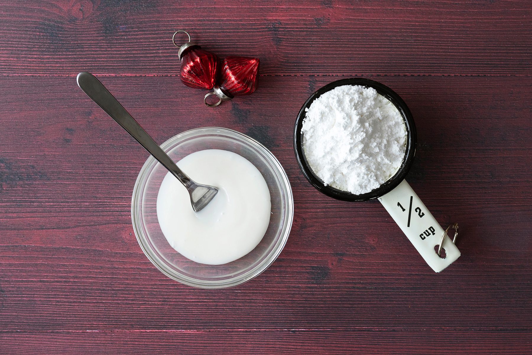 A small glass bowl with white icing and a spoon on a dark red wooden surface, next to a half-cup measuring cup filled with powdered sugar. A red metallic ornament is placed above them.