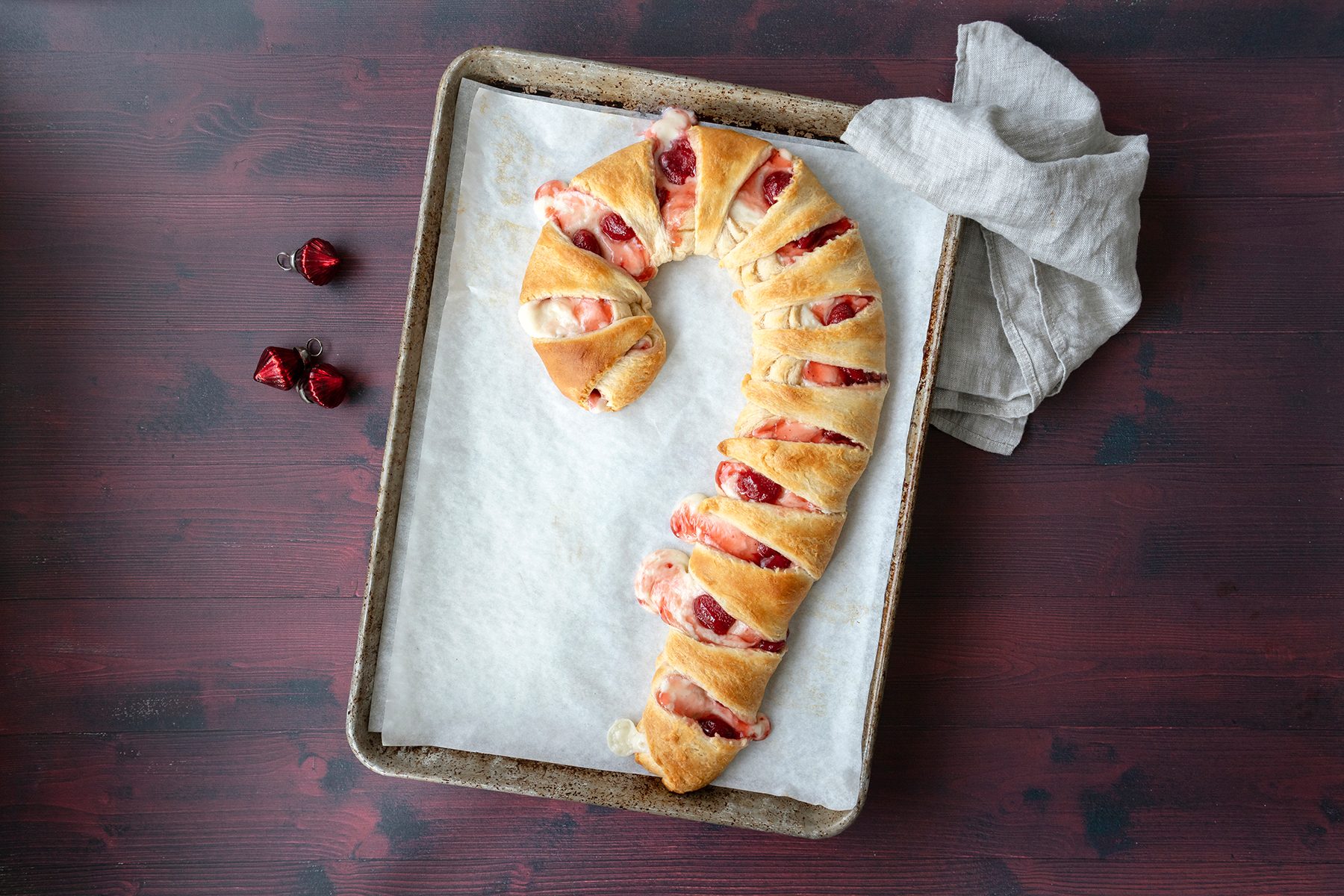 A baked pastry shaped like a candy cane sits on a baking sheet lined with parchment paper. It features layers of golden-brown dough with red and white filling. A few wrapped chocolate candies and a cloth napkin are nearby on a dark wooden surface.