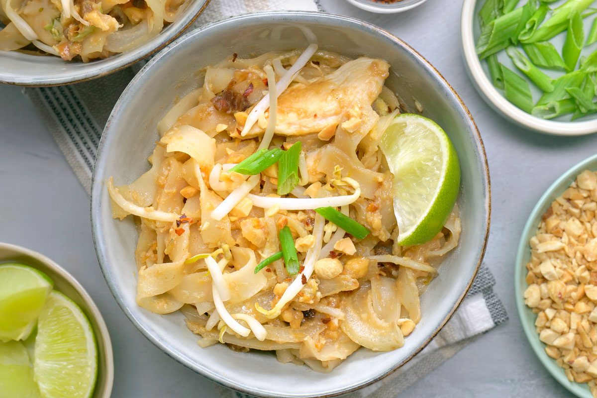 overhead shot of a bowl of Chicken Pad Thai; the noodles are topped with bean sprouts, crushed peanuts, and chopped scallions; a wedge of lime sits on top of the dish; the bowl is sitting on a white linen napkin