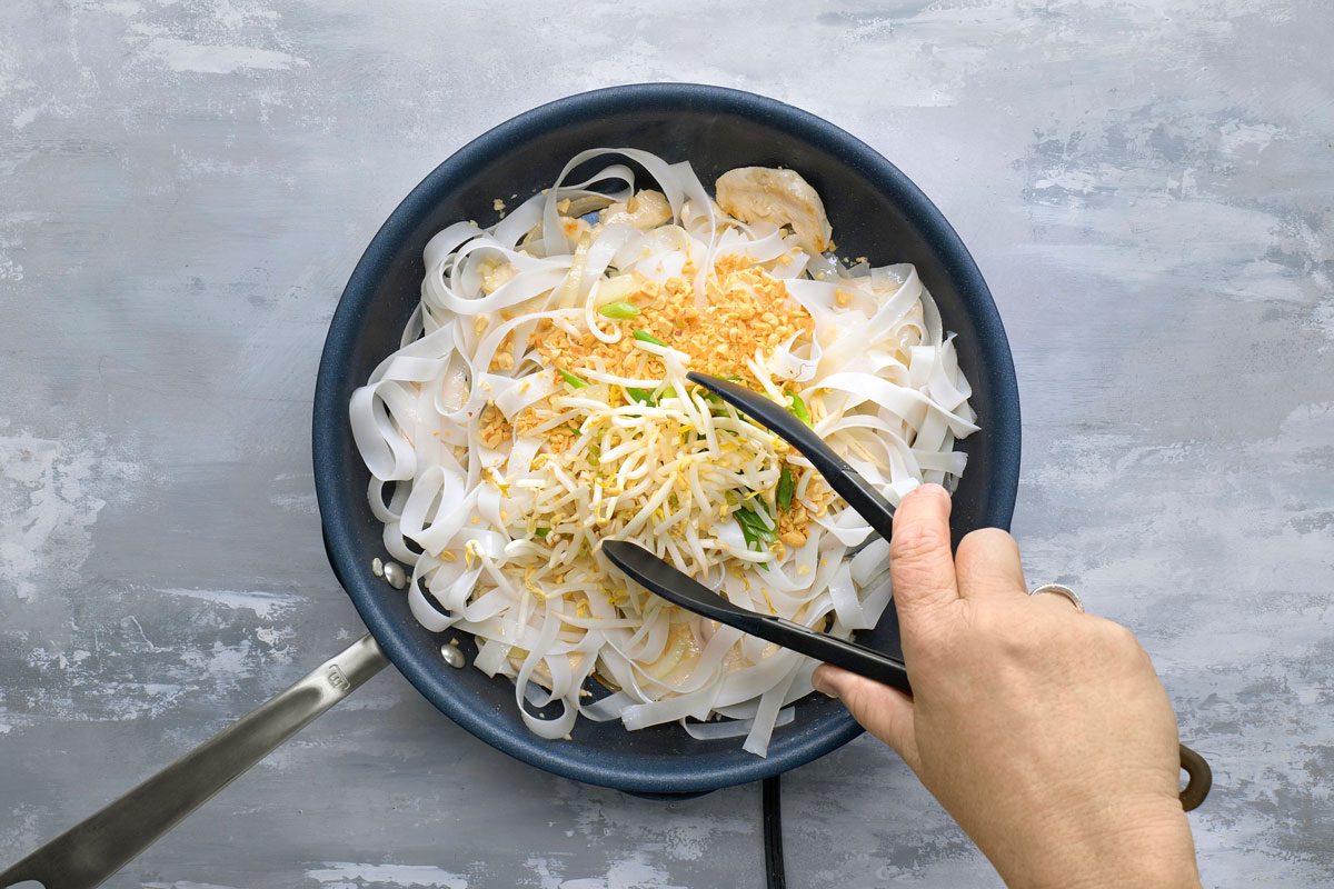 overhead shot of a person cooking pad thai in a black pan over a grey counter; the pan is filled with pad thai noodles, chicken, bean sprouts, and crushed peanuts; the person is using tongs to mix the ingredients together