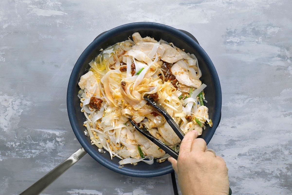 overhead shot of a black frying pan on a light grey surface; the pan contains cooked Pad Thai noodles with chicken, bean sprouts, and green onions; the noodles are white and the chicken is light brown; the noodles are coated in a brown sauce; the pan is on the left side of the image and a hand holding a pair of black tongs is on the right side of the image