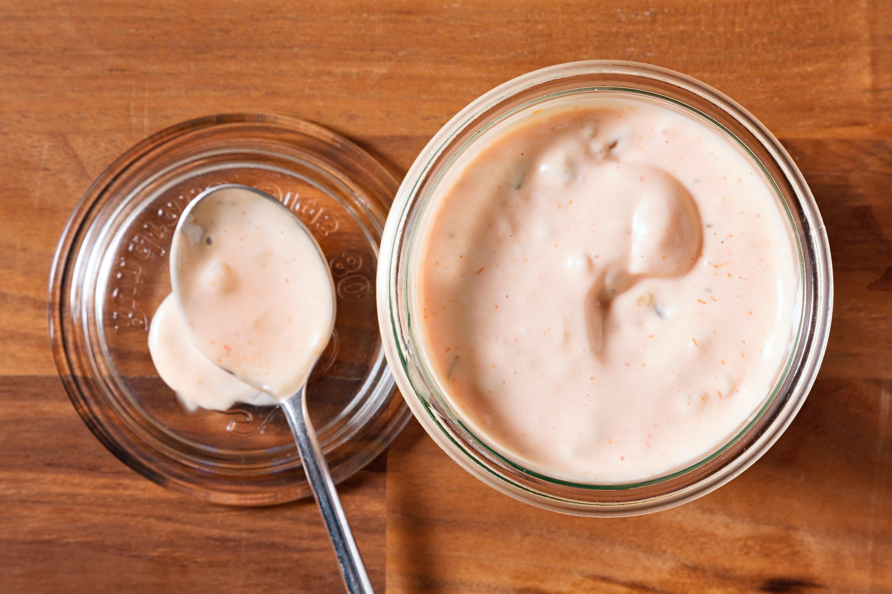 A top-down view of a glass jar filled with creamy Thousand Island dressing on a wooden surface. A spoon rests on the jar's lid beside it, with a dollop of dressing on the spoon.