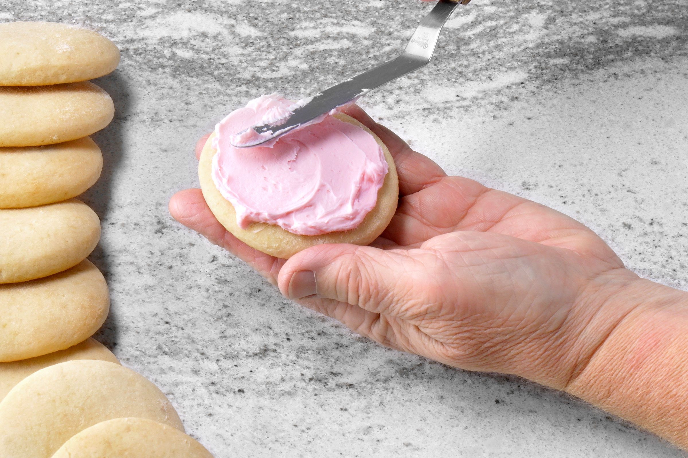Spreading pink colored cookie frosting on baked cookie with a spatula