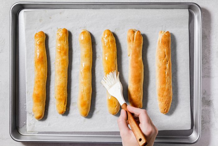 A hand uses a brush to apply butter to a row of six golden-brown breadsticks lined up on a parchment-covered baking sheet.