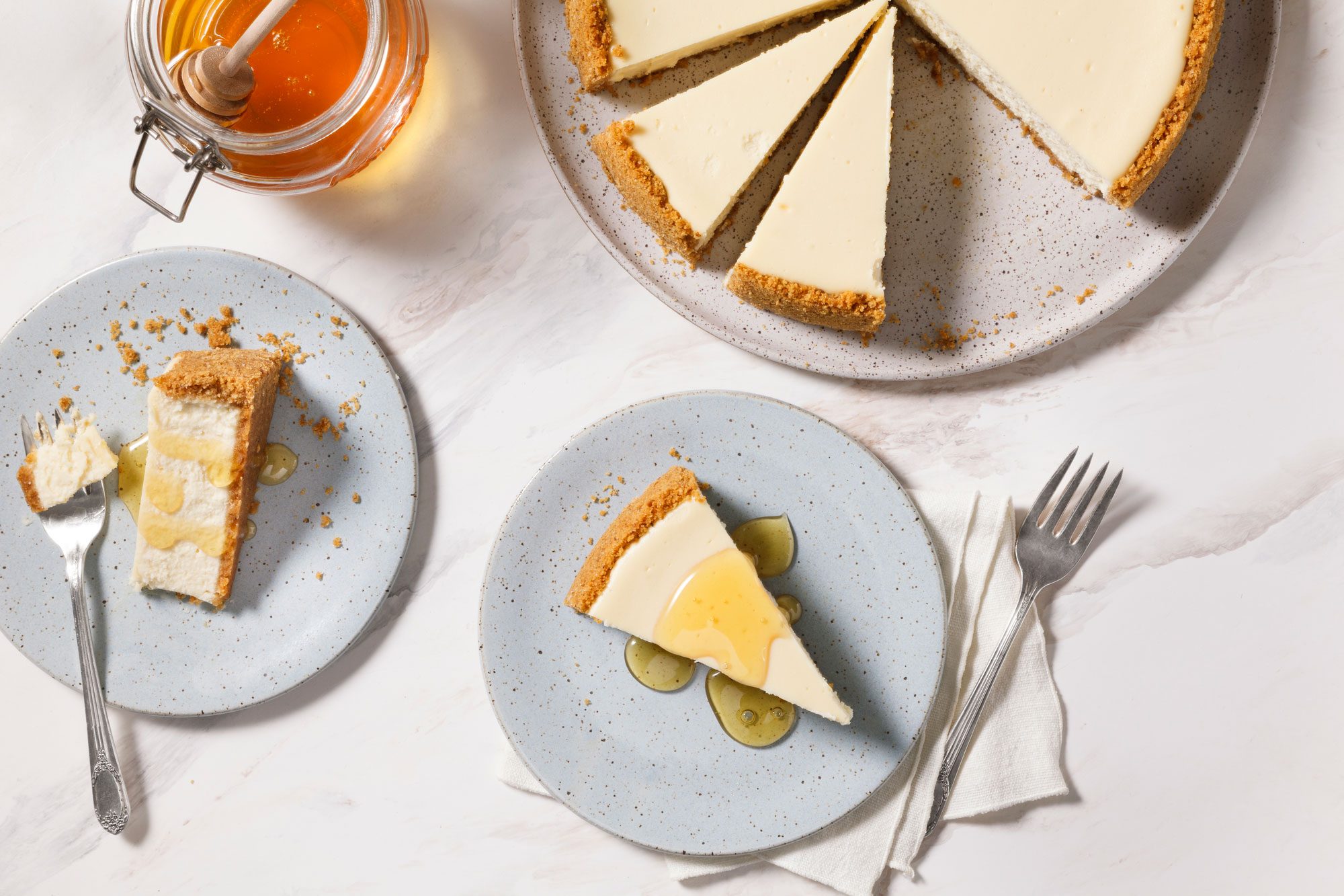 A top-view image of a Goat Cheese Cheesecake on a plate, sliced into pieces. Two servings are on separate plates, one drizzled with honey. A jar of honey with a dipper, a fork, and a napkin are also visible on the table.