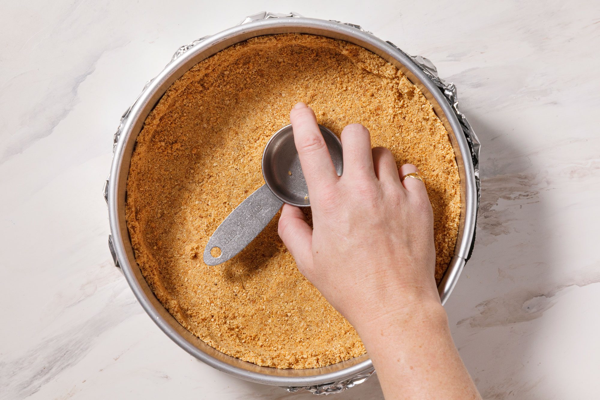 A hand presses a metal measuring cup into a graham cracker crust in a round springform pan on a marble surface.