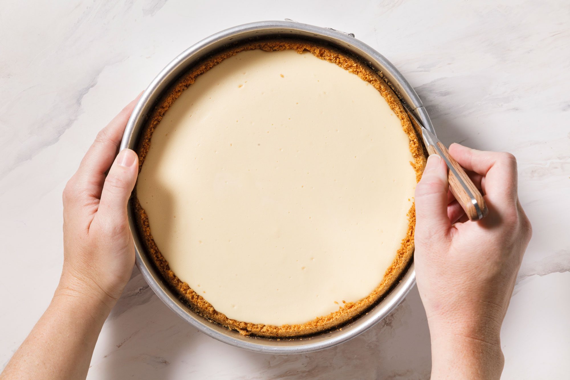A person is holding a round cheesecake in a metal baking pan, with a knife poised to cut it. The cheesecake has a golden crust and a smooth, creamy top.