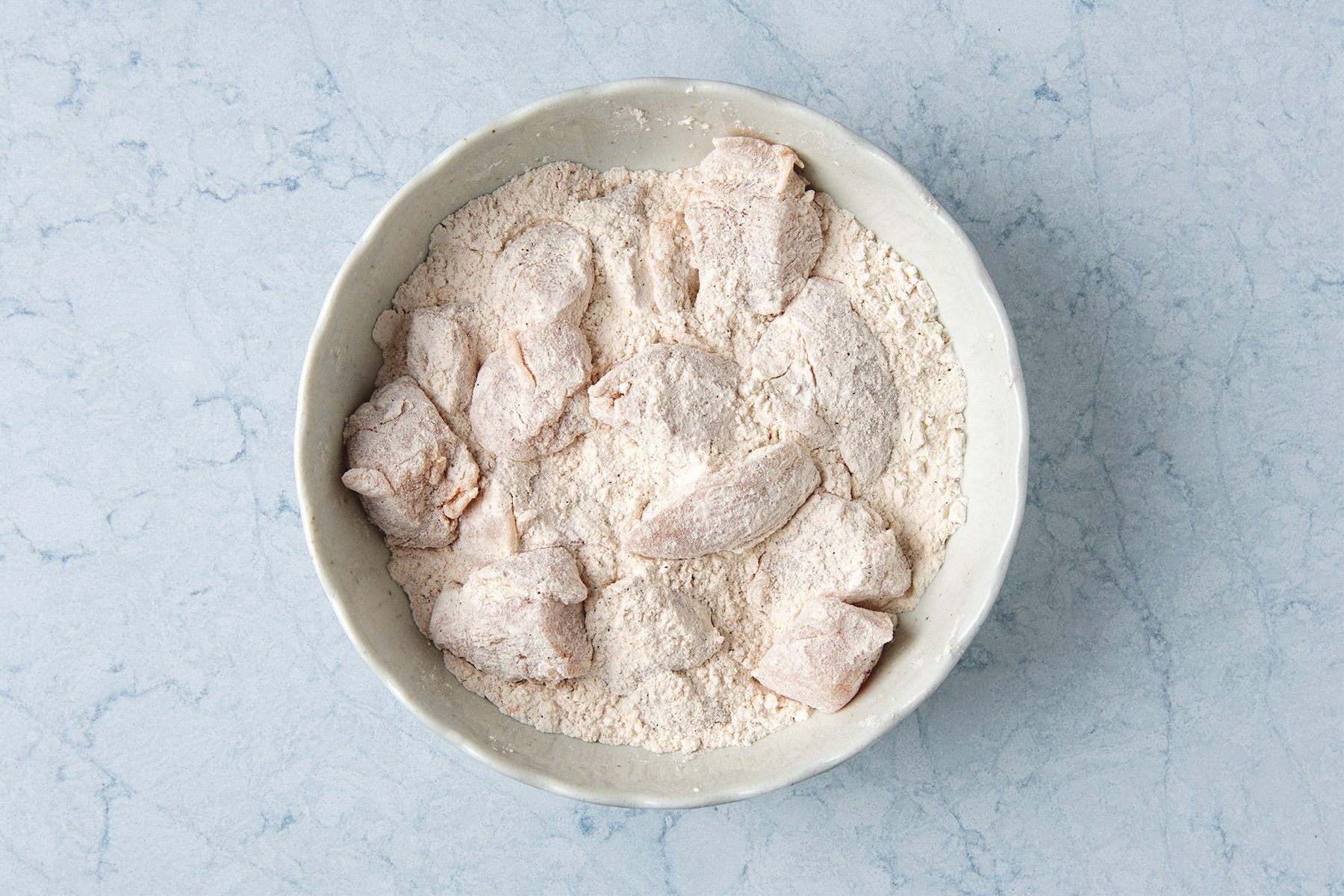 Chicken pieces coated in flour are placed in a white bowl on a light blue marble surface.