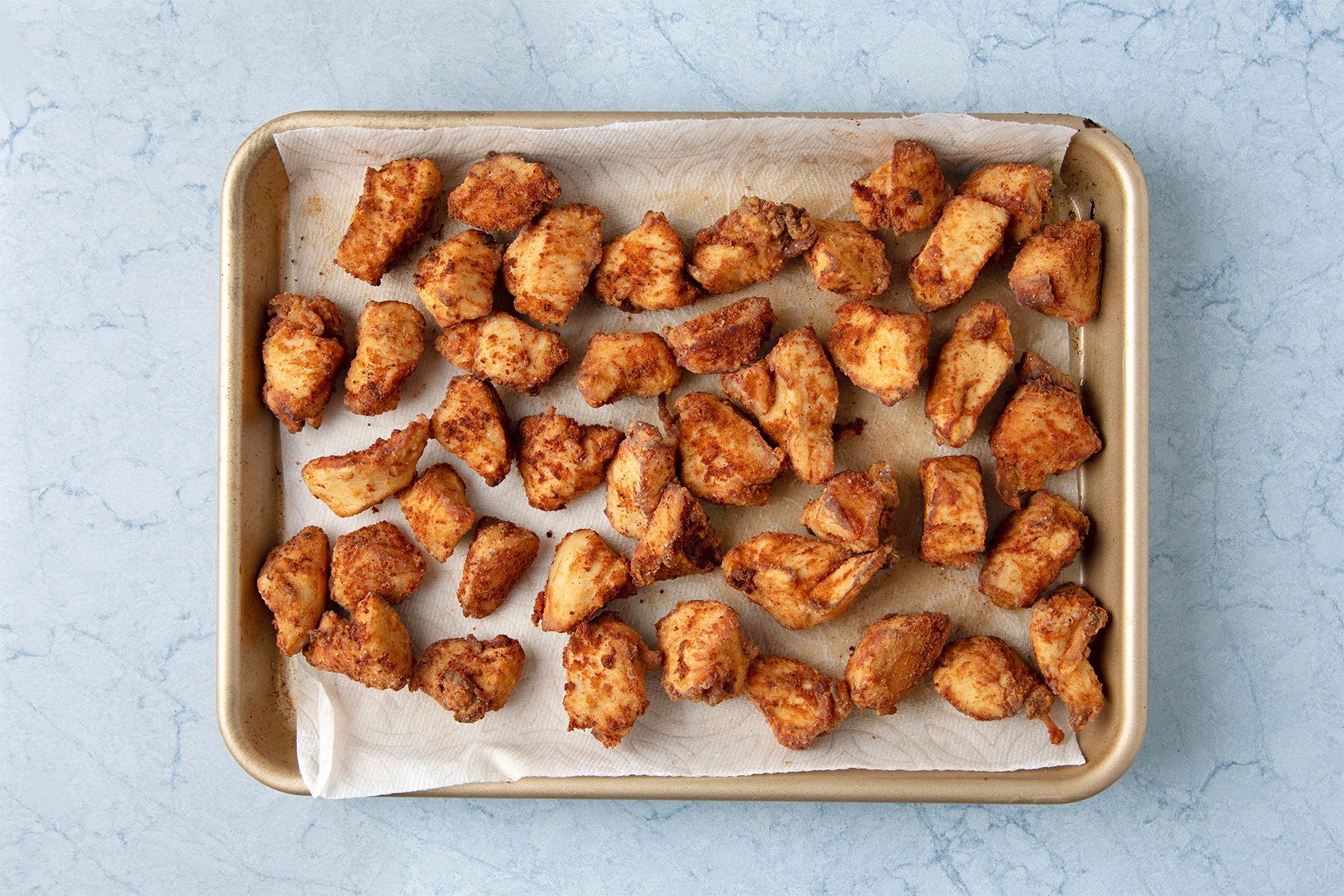 A baking sheet with evenly spaced pieces of fried chicken on parchment paper, set on a light blue surface.